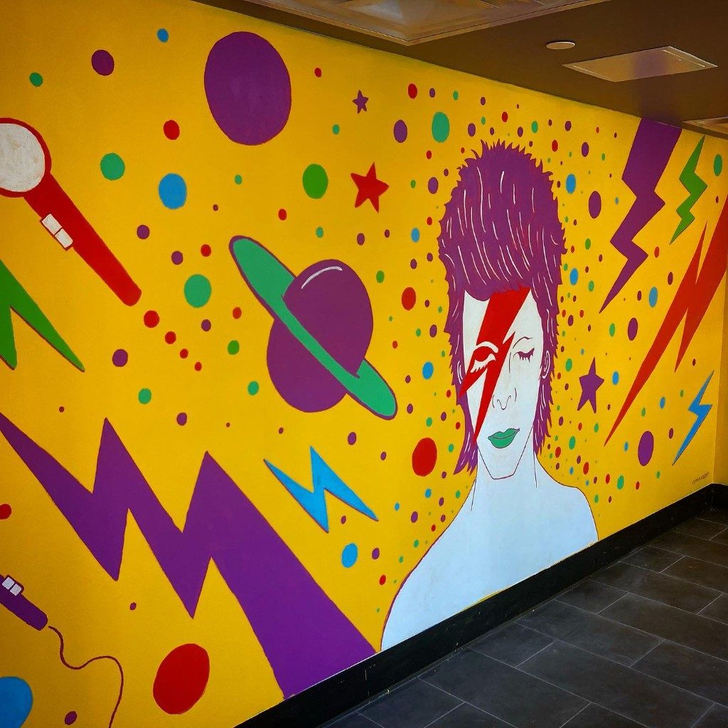 A mural of David Bowie with a red lightning bolt across his face, painted on a yellow wall with lightning bolts, planets, and a microphone.